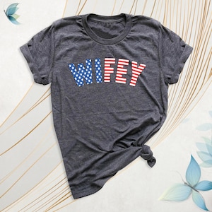 Puede incluir: Camiseta gris oscuro con la palabra "WIFEY" en rojo y azul. El "WIF" est&aacute; en estrellas azules y el "EY" en rayas rojas. La camiseta est&aacute; anudada en la parte inferior.