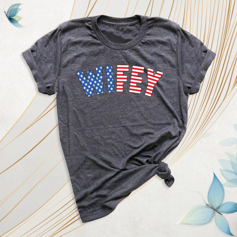 Puede incluir: Camiseta gris oscuro con la palabra "WIFEY" en rojo y azul. El "WIF" est&aacute; en estrellas azules y el "EY" en rayas rojas. La camiseta est&aacute; anudada en la parte inferior.