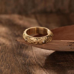 Könnte beinhalten: Ein goldfarbener Ring mit einem aufwendigen Blumenmuster und kleinen, klaren Edelsteinen. Der Ring liegt auf einer braunen, strukturierten Oberfläche, mit einem verschwommenen Holz-Hintergrund. Die detaillierten Schnitzereien und der Kontrast des Goldes erzeugen ein ansprechendes Bild.