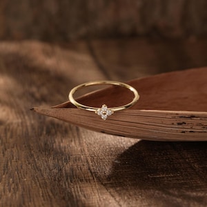 Op de afbeelding: Een delicate gouden ring met een vierbladige bloemvorm, bezet met kleine, heldere edelstenen. De ring ligt op een bruin, getextureerd oppervlak, wat de elegante eenvoud benadrukt. De ring heeft een dunne, gepolijste band.