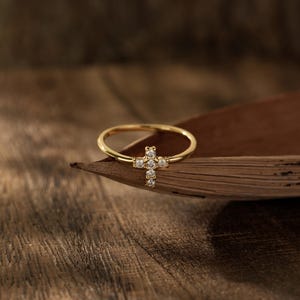 May include: A thin gold ring featuring a small cross-shaped centerpiece adorned with five clear, sparkling stones. The band is smooth and polished, showcasing a delicate design that highlights the cross motif set with brilliant, reflective gems.