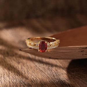 May include: A gold ring featuring a large red oval gemstone surrounded by smaller clear gemstones.