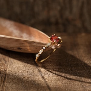 Puede incluir: Un anillo de oro con una piedra preciosa redonda y facetada de color naranja rojizo, montada en cuatro garras. La banda está decorada con una serie de pequeñas piedras redondas y transparentes engastadas en monturas de metal en forma de marquesa.