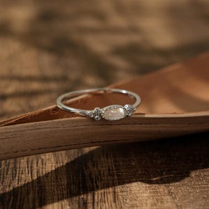 Op de afbeelding: Een delicate zilveren ring met een centrale ovale, lichtgekleurde steen, geflankeerd door kleinere, sprankelende accenten. De ring ligt op een natuurlijk, bruin, getextureerd oppervlak, waardoor een contrast ontstaat tussen het koele metaal en de warme tinten.