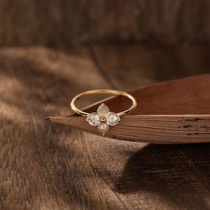 Peut inclure: Une bague en or avec un groupe de pierres précieuses blanches et de perles. La bague est posée sur une surface en bois.