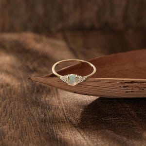 Peut inclure: Une bague en or avec une pierre centrale en opale ovale et de petites pierres d'accent de chaque côté. La bague est présentée sur une surface brune texturée, mettant en valeur son design délicat et son savoir-faire. L'anneau est fin et élégant.