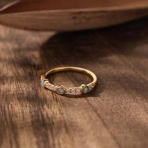 May include: A gold ring featuring small opals and clear stones set in a band. The ring is displayed on a wooden surface. The opals are a light blue colour. The ring is a delicate piece of jewellery.