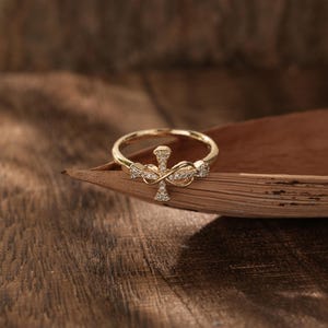 May include: A gold ring featuring a cross design, adorned with small, sparkling stones. The ring sits on a wooden surface, providing a warm, natural backdrop. The cross is the central element of the ring.