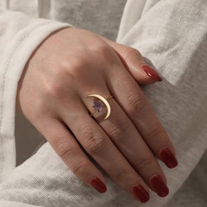 Può includere: Un anello dorato con un design a mezzaluna, con una piccola gemma viola. L'anello è indossato su una mano con smalto rosso, su una camicia bianca a maniche lunghe.