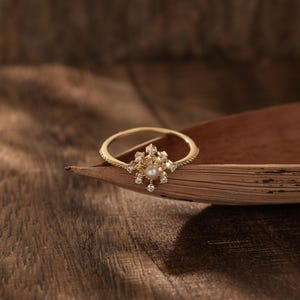 Könnte beinhalten: Ein goldener Ring mit einer Perle und kleinen klaren Steinen, die in einem Blumenmuster eingefasst sind. Der Ring liegt auf einer braunen Holzoberfläche.