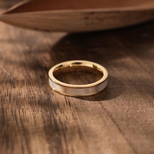 Puede incluir: Un anillo dorado con una incrustación blanca, sobre una superficie de madera oscura. El diseño del anillo presenta una banda dorada con una franja blanca central. Una joya sencilla y elegante.