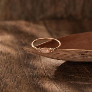 Peut inclure: Une bague en or rose avec une pierre centrale ovale et de petites pierres d'accent de chaque côté. La bague est posée sur une surface naturelle et texturée, mettant en valeur son design élégant et son savoir-faire. La bague est un bijou.