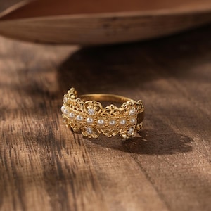Könnte beinhalten: Ein goldener Ring mit einem filigranen Design und einer Reihe weißer Perlen. Der Ring liegt auf einer Holzoberfläche.