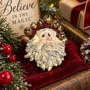 Pode incluir: Um ornamento com o rosto do Pai Natal apresentando uma barba e bigode texturizados na cor creme, bochechas rosadas e uma aba de gorro vermelho escuro adornada com uma fileira de pequenos guizos dourados metálicos e detalhes pintados.