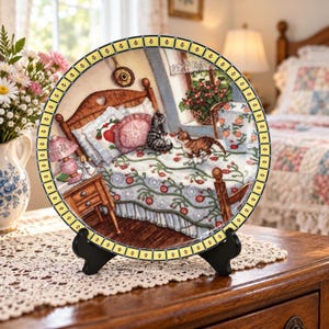 May include: A round decorative ceramic plate featuring a detailed illustration of a bedroom scene with two cats on a bed, a wooden nightstand, and a lamp. The plate has a yellow border with small red flower motifs and is displayed on a black stand.