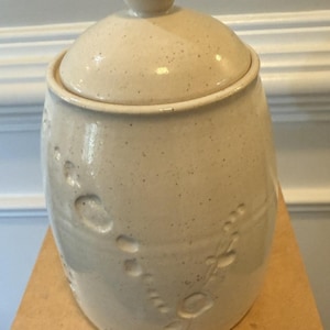May include: A ceramic jar with a lid, in a speckled off-white color. The jar has a rounded shape with a lid and a small knob on top. The jar's surface features a textured design of circular and floral patterns.