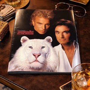 Puede incluir: Una portada de revista con Siegfried & Roy y un tigre blanco. La portada tiene el texto "Siegfried & Roy at the Mirage". La imagen está sobre una superficie oscura con una vela, una bebida y otros elementos decorativos.