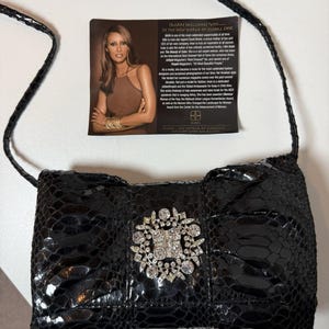 Könnte beinhalten: Eine schwarze Handtasche mit Schlangenleder-Textur, einem dünnen Schulterriemen und einer großen, runden Brosche aus klaren Strasssteinen in der Mitte. Beiliegend ist eine rechteckige Informationskarte mit einem Foto von Iman und Text zu ihrer Karriere.