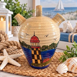 May include: A beige ceramic vessel with two spouts and a central handle, featuring a hand-painted scene of a lighthouse with a red roof and black and yellow checkered base, set against a blue water background with small black birds in the sky.