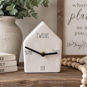 May include: A white ceramic house-shaped clock with black hands and black text indicating the hours twelve, three, six, and nine. The clock features a smooth finish and a central brass-colored mechanism connecting the two black clock hands.