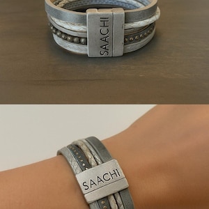 May include: A multi-strand bracelet with a magnetic clasp. The bracelet features gray, white, and silver bands with decorative beads and textured patterns. The clasp is rectangular and has the word "SAACHI" engraved on it.
