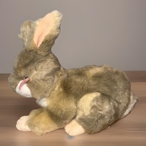 May include: A plush rabbit toy in shades of brown, tan, and white. The rabbit has long ears with pink inner ears, a pink nose, and pink paws. The toy is lying down on a wooden surface.