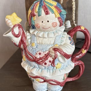 May include: A whimsical ceramic teapot in the shape of a doll. The doll has a colorful patchwork hair, rosy cheeks, and is wearing a blue and white dress. The teapot features a star-shaped spout and a red handle with yellow stars. Ribbons and floral details adorn the teapot.