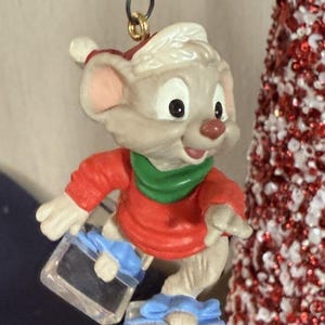 Peut inclure: Ornement de Noël représentant une souris de dessin animé portant un bonnet de Père Noël, une chemise rouge et une écharpe verte. La souris tient deux boîtes cadeaux avec des nœuds bleus. Un petit sapin de Noël décoré est en arrière-plan.