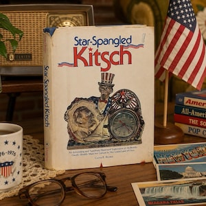 以下が含まれることがあります： カーティス・F・ブラウン著のハードカバー本『Star-Spangled Kitsch』。表紙にはアンクル・サムと時計のイラストが描かれています。本の周りには、1776-1976のエンブレムが入った白いマグカップ、茶色の眼鏡、小さなアメリカ国旗、絵葉書が置かれています。