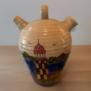 May include: A ceramic jug with three spouts and a handle. The jug is painted with a lighthouse scene, featuring a red-domed lighthouse with a black and yellow checkered pattern. The background includes blue water, green trees, and flying birds.