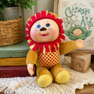 May include: A Cabbage Patch Kids doll dressed in a lion costume with a red and yellow felt mane, blue eyes, and a tan plush body featuring a red polka-dot pattern on the chest. The doll has a soft vinyl face with painted whiskers and a pink mouth.