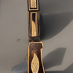 Peut inclure: Un couteau en bois foncé avec des sculptures complexes et une incrustation de couleur claire. Le manche présente un motif en forme de feuille, tandis que la lame a un motif géométrique. Le couteau est probablement un objet décoratif ou de cérémonie.