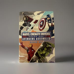 Puede incluir: Un set en caja con Marvel Cinematic Universe Fase Uno: Avengers Assembled. El arte de la caja muestra ilustraciones de Thor, Capitán América, Iron Man y Hulk. La caja tiene un diseño vintage con el texto del título en un lugar destacado.