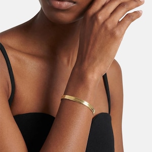 Könnte beinhalten: Ein goldfarbenes, flaches Fischgrät-Armband. Das Armband wird am Handgelenk einer Person getragen. Das Armband hat eine strukturierte Oberfläche und ein klassisches Design. Die Person trägt ein schwarzes Oberteil.