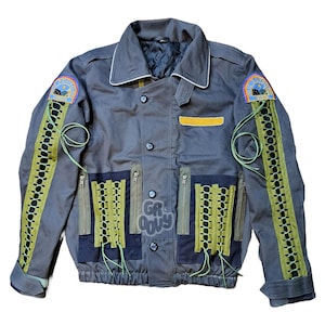 May include: A dark grey utility jacket with green and black lace-up details on the sleeves and front pockets. The jacket has a white collar and two yellow patches on the sleeves.