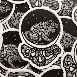 May include: Black and white sticker featuring a toad inside a glass sphere with stars. The toad is sitting on a skeletal hand.