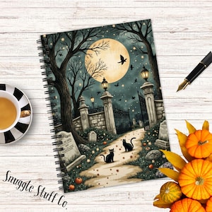 May include: Spiral-bound notebook with a Halloween-themed illustration. The cover features a spooky graveyard scene with black cats, a full moon, and pumpkins. A cup of tea, a pen, and pumpkins are also visible.