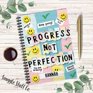 May include: A colorful spiral bound notebook with a motivational quote "Progress not perfection" on the cover. The cover is decorated with ripped paper, smiley faces, and the name "Hannah".