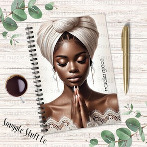 May include: A spiral bound notebook with a white background and a woman with dark skin wearing a white turban and a white lace top. The woman has her hands clasped in prayer. The text "natalia grace" is written vertically on the right side of the notebook.