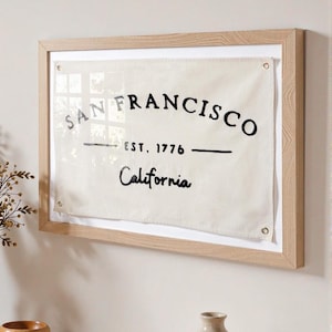 Può includere: Opera d'arte da parete incorniciata su tela. Il testo nero "SAN FRANCISCO EST. 1776 California" è stampato su uno sfondo bianco sporco. La cornice è in legno chiaro. L'opera è appesa a una parete bianca.