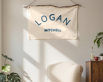 Könnte beinhalten: Ein cremefarbenes Banner mit dem Namen "LOGAN" über "MITCHELL" in marineblauen Buchstaben, das an einem Ast aufgehängt ist. Das Banner hängt über einem cremefarbenen Sessel. Topfpflanzen und ein Holzschrank sind ebenfalls zu sehen.