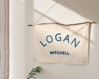 Könnte beinhalten: Cremefarbenes Banner mit dem Namen "LOGAN" über "MITCHELL" in blauer Schrift. Das Banner wird von einem natürlichen Holzstab und einer weißen Schnur gehalten. Es hängt an einer weißen Wand, mit einer Pflanze im Vordergrund.