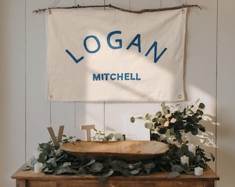 Könnte beinhalten: Ein cremefarbenes Banner mit dem Namen "LOGAN" und dem Nachnamen "MITCHELL" in blauen Buchstaben, aufgehängt an einem Ast. Darunter ein Holztisch mit einer Holzschale, Grünzeug, Kerzen und Holzbuchstaben.