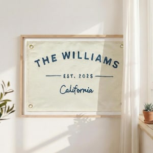 Op de afbeelding: Een rechthoekige wandbanner van gebroken wit canvas met donkerblauw geborduurde tekst die THE WILLIAMS, EST. 2025 en California leest. De banner is ingelijst in licht hout en heeft vier messingkleurige metalen ringen in de hoeken.