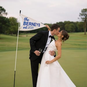 Könnte beinhalten: Ein küssendes Paar auf einem Golfplatz, mit einer weißen Fahne mit der Aufschrift "the BERNEYS est. 2025" in blauer Schrift. Die Braut trägt ein weißes Hochzeitskleid, der Bräutigam einen schwarzen Smoking. Der Hintergrund zeigt grünes Gras und Bäume.