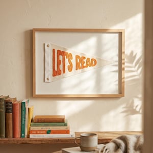 Könnte beinhalten: Ein gerahmtes Wimpel mit den Worten "LET'S READ" in orangefarbener Stickerei. Der weiße, dreieckige Wimpel ist in einem hellen Holzrahmen montiert. Das Bild wird über einem Regal mit Büchern und einer Tasse angezeigt.