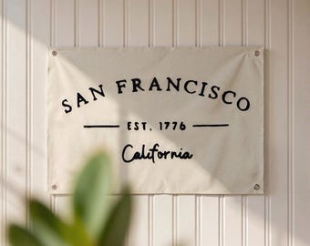 Może przedstawiać: Prostokątny baner z kremowego płótna z napisem "SAN FRANCISCO" w zakrzywionej czcionce, poniżej "EST. 1776" i "California" w czcionce skryptowej, wszystko w kolorze czarnym. Przymocowany do białej ściany z pionowymi panelami.
