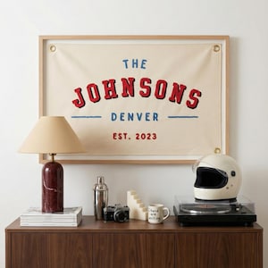 Puede incluir: Un letrero de lona enmarcado con el texto "THE JOHNSONS DENVER EST. 2023" en rojo y azul. Una lámpara de mesa, una coctelera, una cámara, una vela, una taza, un casco y un tocadiscos están sobre un mueble de madera.