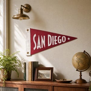 以下が含まれることがあります： 濃い赤色のフェルト製三角形ペナントで、白い文字で「SAN DIEGO」と書かれ、小さな白い星が添えられています。ペナントの左側には白い縦帯があり、吊り下げるための2つの金属製ハトメが付いており、壁に飾られています。