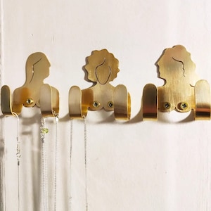 May include: Three gold metal wall hooks shaped like female figures with two hooks on each. The hooks are mounted on a white wall and have necklaces hanging from them.